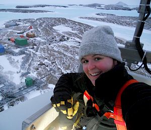 A selfie of a woman wearing a beanie and harness, on a high surface looking down towards station buildings on the ground.