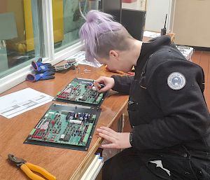 Woman with dyed purple hair sits at desk soldering electronics circuit boards.