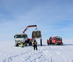 Expeditioners load a pallet of cargo onto a tracked crane parked nearby on ice, next to a smaller red tracked vehicle..