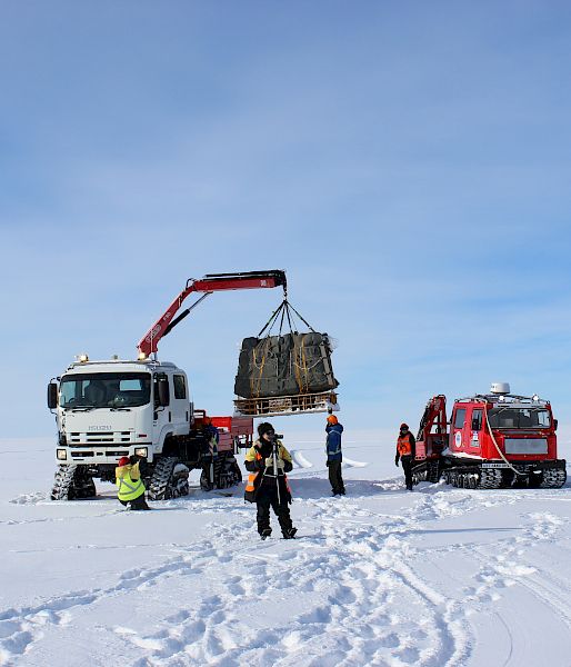 Expeditioners load a pallet of cargo onto a tracked crane parked nearby on ice, next to a smaller red tracked vehicle..