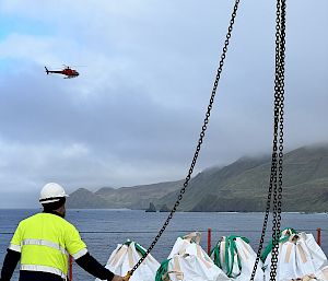 A helicopter flies over the water between a ship and an island. An expeditioner stands on the deck of the ship, preparing large sacks of cargo to be lifted by the helicopter.