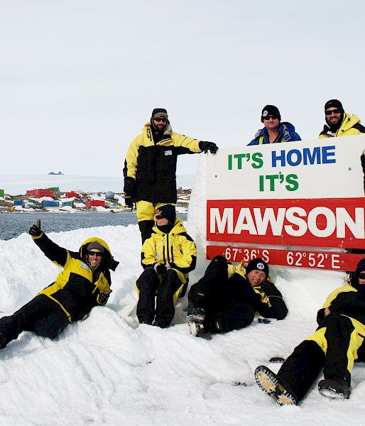 Expeditioners posing with the Mawson sign on West Arm. Station in background