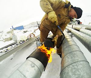 Man outdoors using flaming torch to apply heat shrink to a join in a large silver pipe.