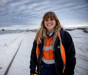 An expeditioner in high-vis stands on the ice. A large orange and white ship is in the distance behind her.
