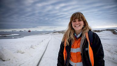 An expeditioner in high-vis stands on the ice. A large orange and white ship is in the distance behind her.