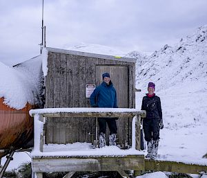 Two people stand on the wooden deck of a hut on a snowy hillside.