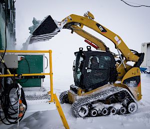 Expeditioner in small yellow digger empties snow from the shovel into a large container attached to side of green building