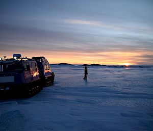 Expeditioner stands with a tracked vehicle on the ice, and the sunset in the distance