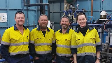 Four people in high visibility clothing in workshop, smiling at camera.