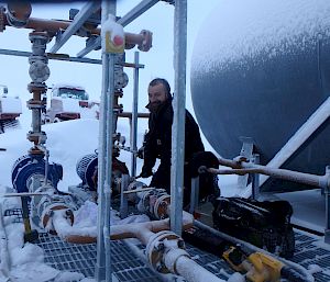 An expeditioner kneeling beside a series of pipes next to a large cylindrical tank. The pipes and ground around him are covered in snow.