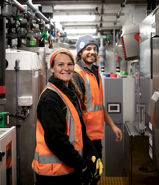 2 expeditioners wearing hi-vis stand smiling in a room of metallic equipment.