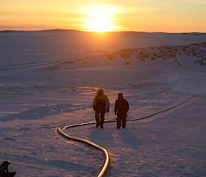 Two men walking centre picture along a large fuel hose which is lying along the snow, in the distance rocky ridges with the sun just rising on the horizon
