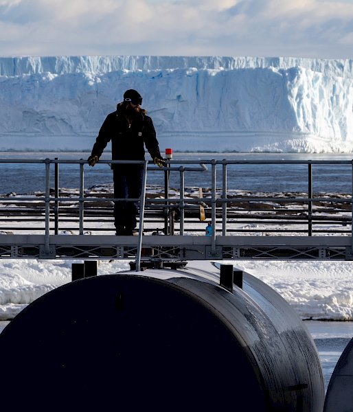 An expeditioner stands on top of a fuel tank with a beautiful landscape in the background - dramatic ice cliffs and a bay.
