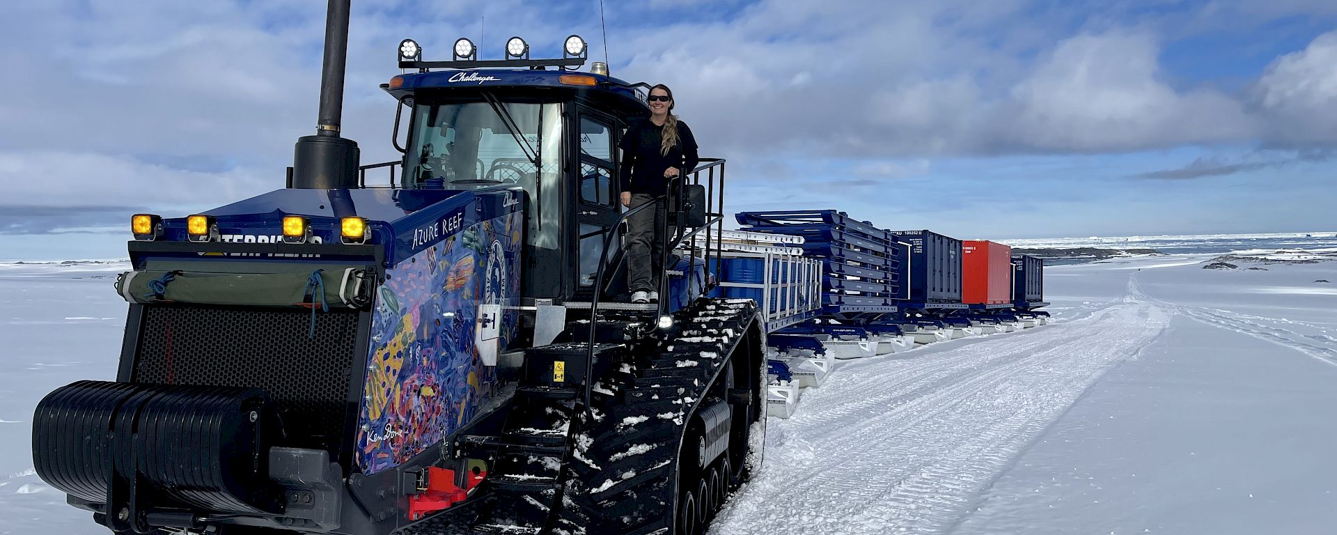 Expeditioner stands on a tractor on the ice, which is pulling several containers and sleds behind.
