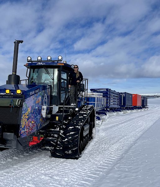 Expeditioner stands on a tractor on the ice, which is pulling several containers and sleds behind.