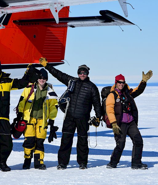 Four happily waving expeditioners, near the tail of the aircraft, on the white ski landing area