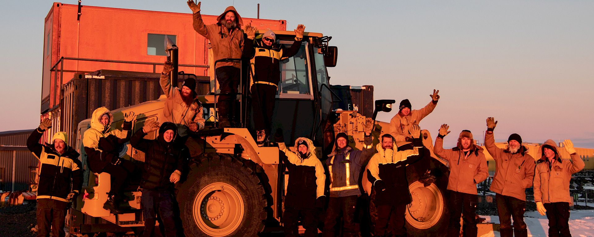 Group of expeditioners standing on and near a large loader truck, waving at the camera at sunset.
