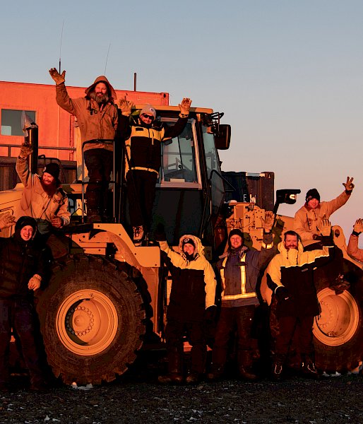Group of expeditioners standing on and near a large loader truck, waving at the camera at sunset.