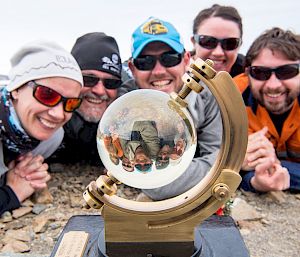 Expeditioners pose around the glass ball of the sunshine recorder that is giving an inverted reflection of their faces.