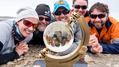 Expeditioners pose around the glass ball of the sunshine recorder that is giving an inverted reflection of their faces.