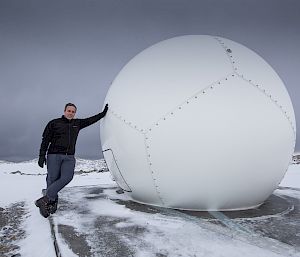 Man in black jumber and grey pants on left, leaning against a large white dome in centre picture. On icy concrete ground with grey sky overhead.