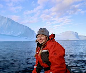 An expeditioner on a boat poses for the camera in front of an impressive iceberg.