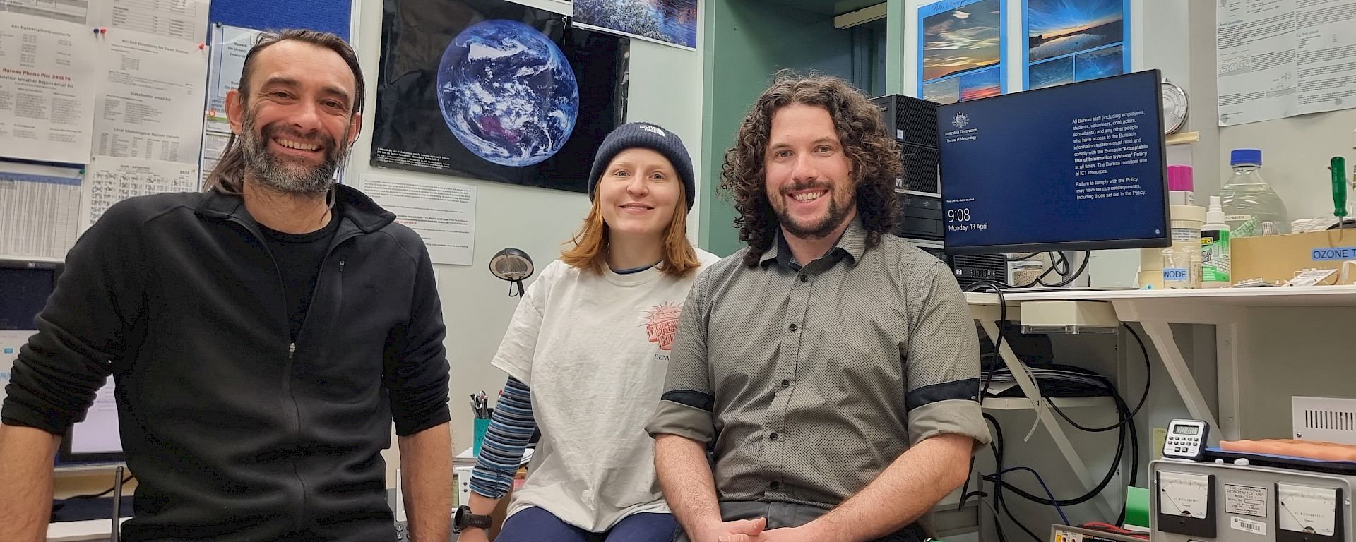 3 expeditioners sit in an office with images and a computer screen showing weather data in the background.