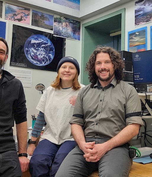 3 expeditioners sit in an office with images and a computer screen showing weather data in the background.