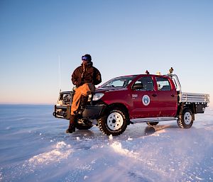 Woman in cold weather gear sitting on bonnet of red ute on ice.