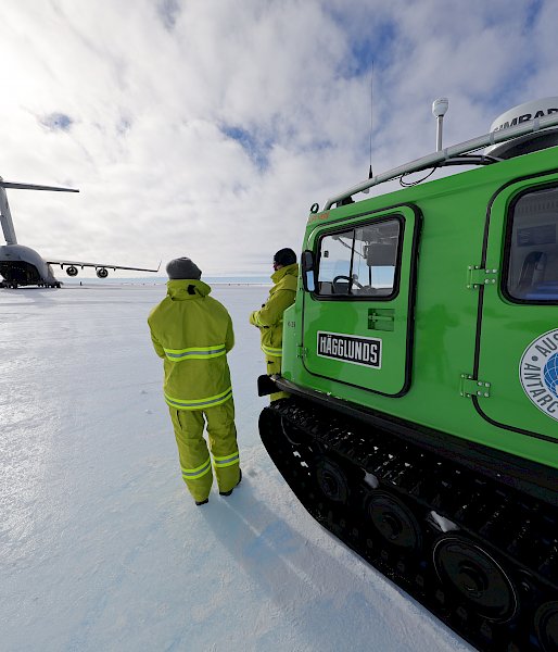 A large aircraft sits on the ice under a blue sky. 2 expeditioners stand next to a green vehicle in the foreground.