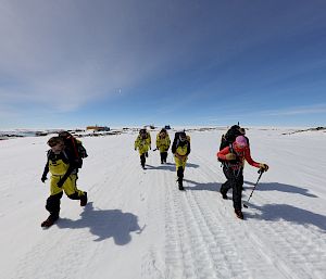 A group of expeditioners walk through an icy landscape under a clear, blue sky.