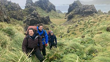 A small group of expeditioners walk through a lush, green landscape with steep rock formations. There is a bay in the distance.