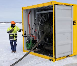 Expeditioner standing next to container on ice monitoring deployment of fuel line from hose reel