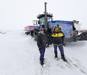 Two expeditioners stand on the ice in front of a tractor adorned with blue-toned artwork.