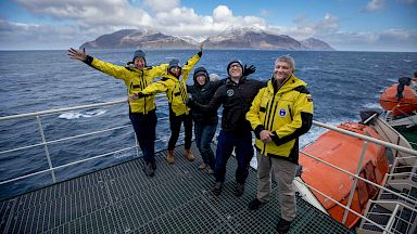 5 people stand on the deck of a ship with their arms outstretched. behind them there is an island on the horizon.