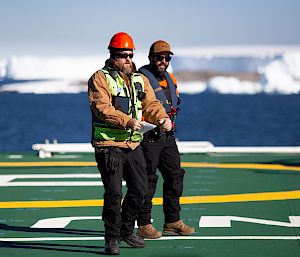 2 expeditioners on the deck of a ship. In the background, icebergs float in blue water.