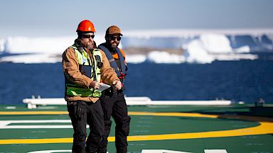 2 expeditioners on the deck of a ship. In the background, icebergs float in blue water.