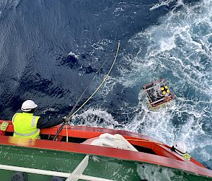 A yellow instrument floats in the ocean. A crew member leans out of an orange ship and pulls a rope.