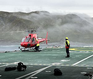 A red helicopter on the green deck of a ship. There are cloudy mountains in the background.