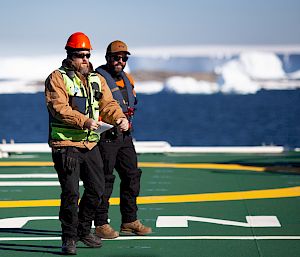 2 expeditioners on the deck of a ship. In the background, icebergs float in blue water.
