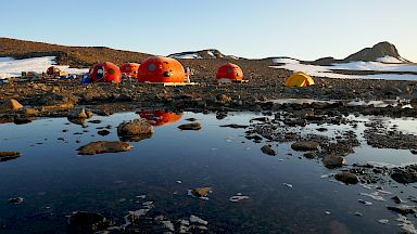 Four orange melon shaped structures behind a lake, with their reflection appearing in the lake water. Two yellow tents pitched behind them, with snow-covered rocky hills in the background.