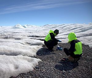 Two people crouched over collecting rock samples in what looks like a sea of cloud, but it's actually snow and ice