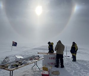 Group of expeditioners with ice core drill at field camp with sun overhead