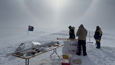 Group of expeditioners with ice core drill at field camp with sun overhead