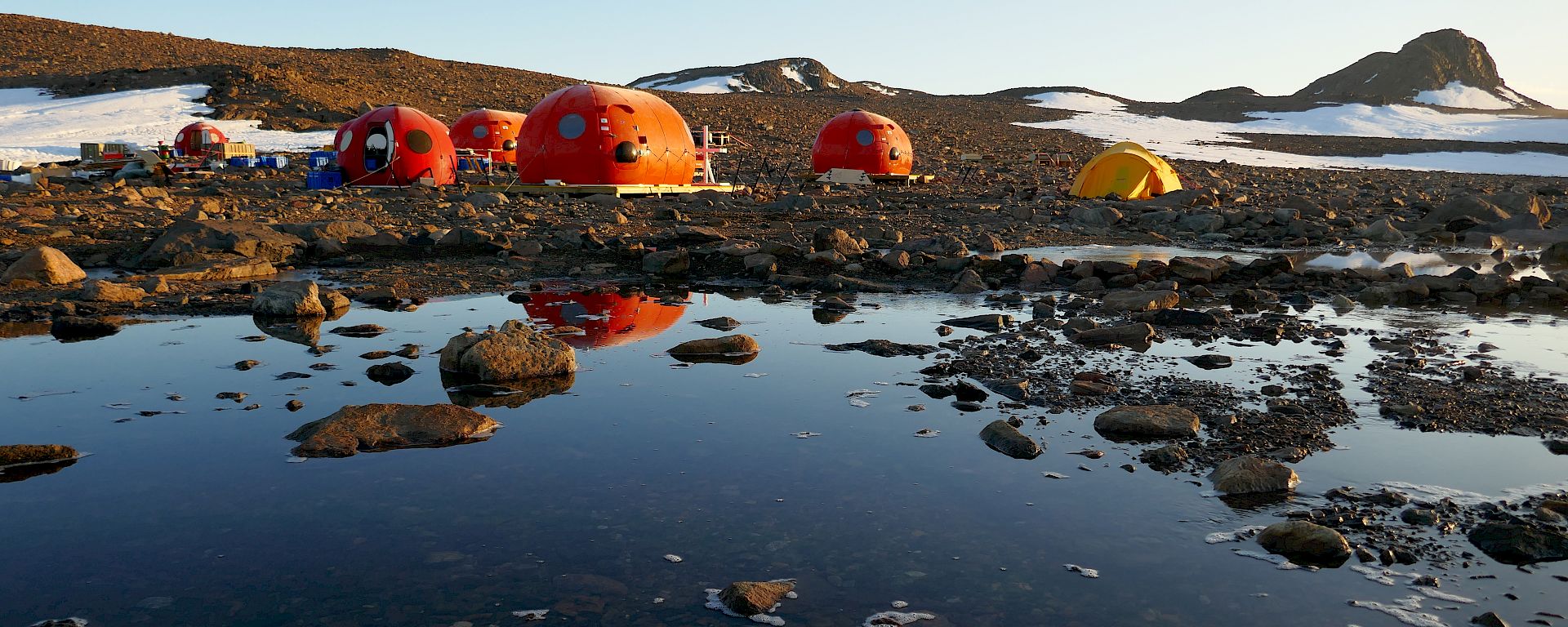 Four orange melon shaped structures behind a lake, with their reflection appearing in the lake water. Two yellow tents pitched behind them, with snow-covered rocky hills in the background.