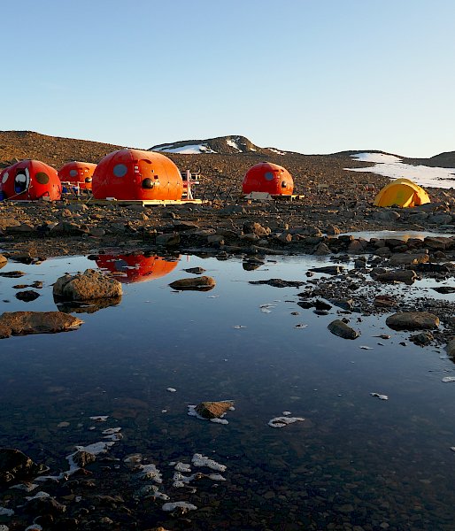 Four orange melon shaped structures behind a lake, with their reflection appearing in the lake water. Two yellow tents pitched behind them, with snow-covered rocky hills in the background.