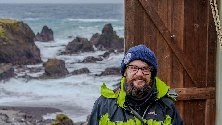 Andy standing near a door, with the Macquarie Island shore in the background