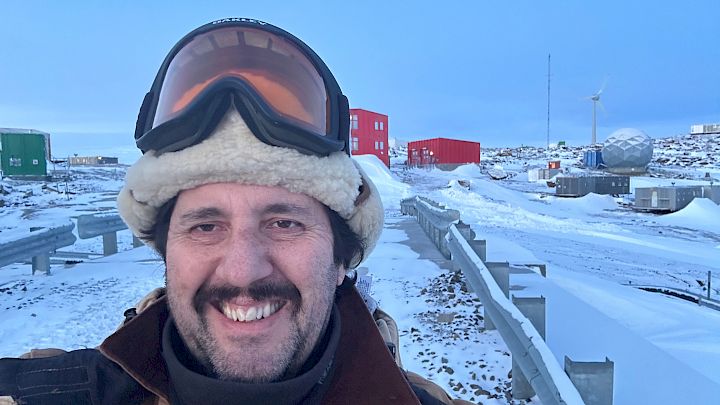 Expeditioner at Mawson station with some station piping and infratsructure behind him covered in snow