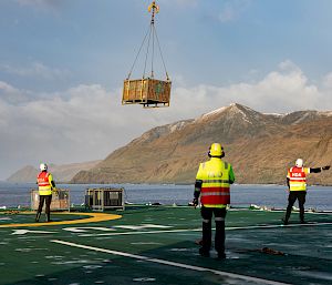 A helicopter is carrying a sling load of the deck of RSV Nuyina. Several people are standing on the deck ensuring the load is correctly picked up.