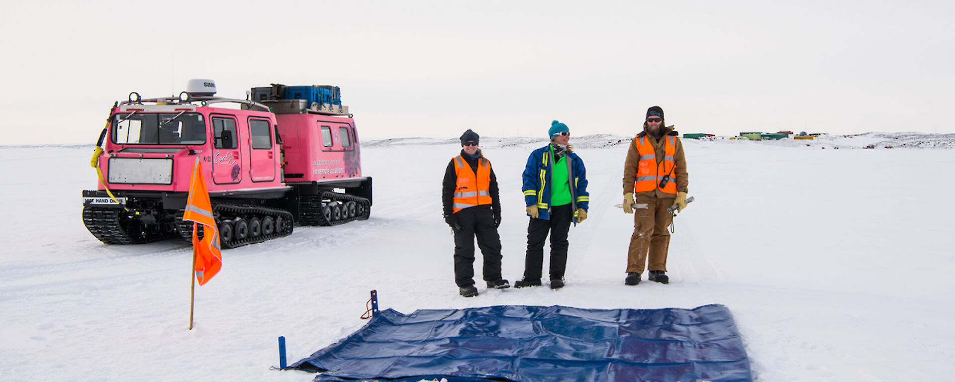 Expeditioners standing near marked out area on sea ice with snow pole line marker and flag with oversnow vehicle behind